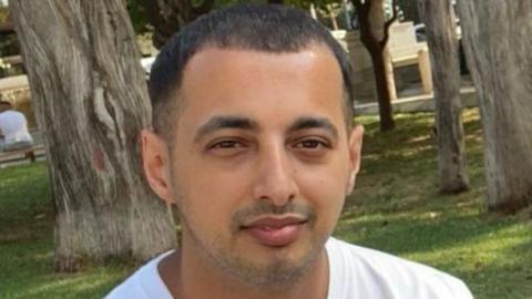 A man in his 30s wearing a white T-shirt. Trees and grass are behind him.