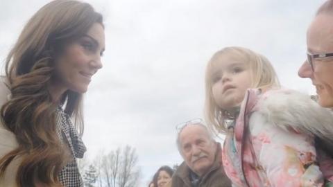 Kate looking at a little blonde-haired girl who is being held by a woman wearing glasses and is wearing a pink jacket with a fur hood