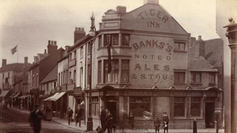 Black and white archive picture of a pub, with Tiger Inn painted on the eaves, under which further painting reads "Banks's noted ales and stouts". People mill around on foot on the streets outside.