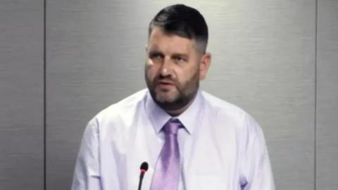 Alan Paton, a bearded man in a purple shirt and tie is sitting in front of a grey wall.