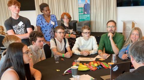 Young people are sitting around a round table looking at a man who is teaching the class. He is wearing a green shirt and glasses.