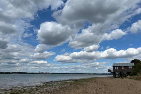 Coastal shoreline with sailboats, a stilted wooden building, and dramatic cloudy sky in Wrabness, Essex