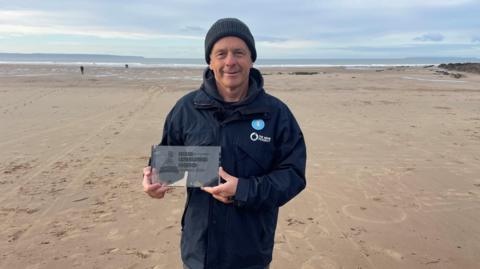 Ian Bennett stood on Croyde beach in North Devon on an overcast day, wearing a coat and woolly hat - holding a glass 'Unsung Hero' award. He is stood on the sand with the sea in the background.