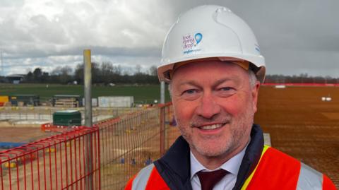 Steve Reed is seen standing in the middle of the picture smiling at the camera. He has a white hard hat on that says Anglian Water in blue writing at the front. He is wearing a red and grey hi-vis jacket, and a white shirt and red tie. 