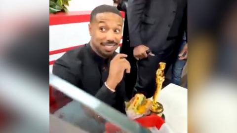 Michael B Jordan smiles at an In-N-Out Burger restaurant as he holds a burger. His Oscar sits on the table next to him.