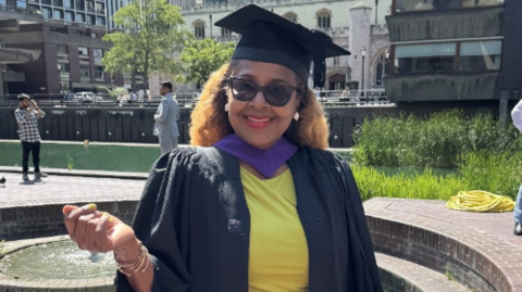 Angella Callam in her graduation garbs, wearing sunglasses, a yellow top underneath her graduation gown. Behind her is a concreted paving area and some greenery. She wears red lipstick and has light brown hair that falls behind her shoulders.