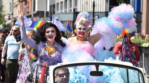 Two participants in the 2023 Belfast Pride parade in an open top car in Belfast City Centre. They are wearing pink and purple dresses, one has long black hair and the other has blue and pink hair pulled high on top of their head.