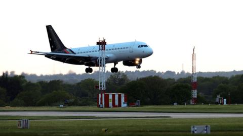 A plane with a white body and blue tail with white stars is seen landing from the side on a runway against a bright sky.