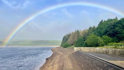 Three quarters of a rainbow over the sea and trees to the right, with a beach in the distance, under a powdery blue sky