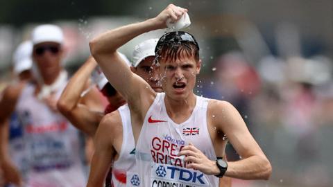 An athlete in a white vest top with Great Britain written on it in red is pouring a sponge full of water over his head. He has dark hair and black sunglasses on top of his head. There are other athletes just visible behind him