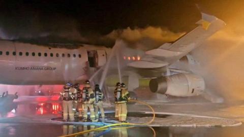 Six firefighters on an airport runway spray hoses onto the side of a jet airplane at night