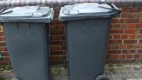 Two grey wheelie bins in front of a brick wall.