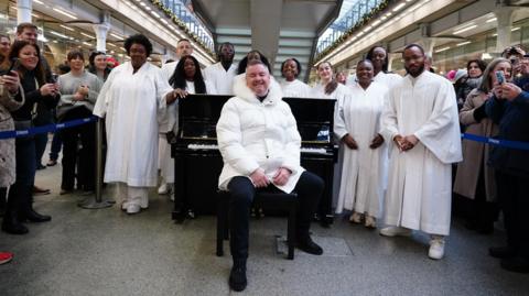 Tony Mortimer is sat at a black piano smiling at the camera, wearing a white puffer jacket with a furry white hood and surrounded by 10 members of the London Gospel Choir wearing white robes. A number of commuters are in the background smiling and holding their phones