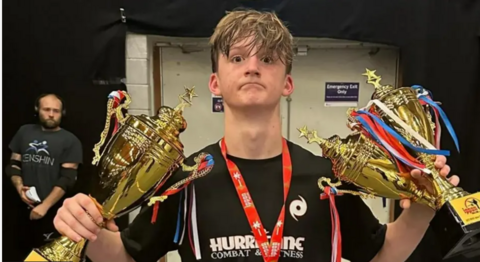 Alex Eastwood, 15, is holding two trophies and wearing medals around his neck.