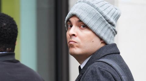 Che Homersham entering Southwark Crown Court wearing a charcoal jacket, grey beanie and with a black rucksack over one shoulder.