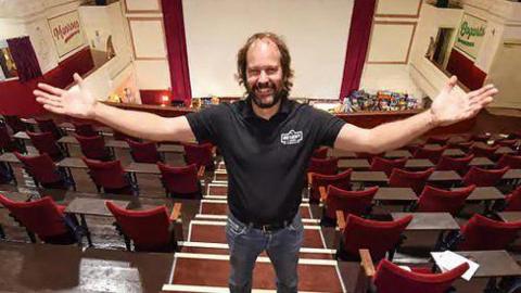 Rick Taylor with long brown hair and beard wearing black t-shirt and jeans stands at the top of tiered red velvet seats in the centre aisle of a cinema with the cinema screen behind him. He is holding his hands out and smiling. 