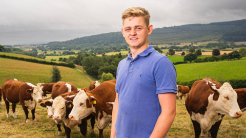 A blonde man in a blue T-shirt, is standing in a field with brown and white cows in the background. Behind them are rolling hills with a lot of green trees visible in the distance.