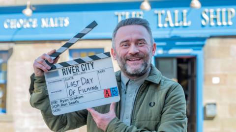 Stephen Purdon stands outdoors holding a film clapperboard that reads “River City – Last Day of Filming.”
The background shows a blue-painted building with signage for “The Tall Ship.”
