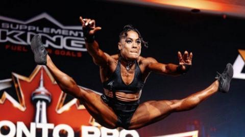 A woman does the splits in mid air, dressed in a black crop top and pants, in front of a colourful backdrop .