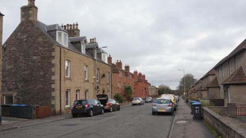 A suburban street in Galashiels with houses up both sides and cars parked on both sides of the road as well