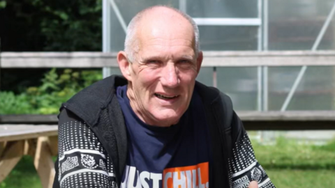 Roy sitting on a wooden bench- wearing a black sleeveless jacket and a navy full-sleeve T-shirt 