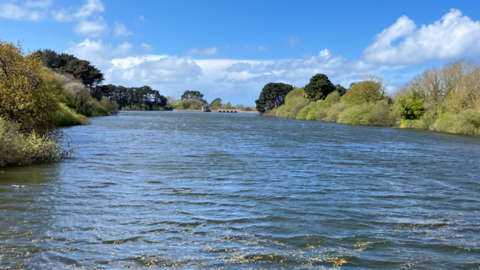 A picture of St Saviour's Reservoir. There is a large body of water with trees either side and a bridge in the distance.