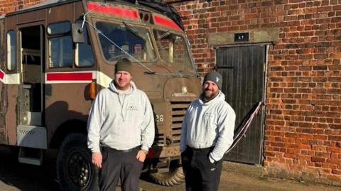 Two men standing in front of a military-style vehicle. It is painted a brown camouflage pattern with red and white accents. Both men are dressed in light grey hoodies, dark trousers and black shoes. They are both wearing beanies. In the background is a brick wall with a wooden door.