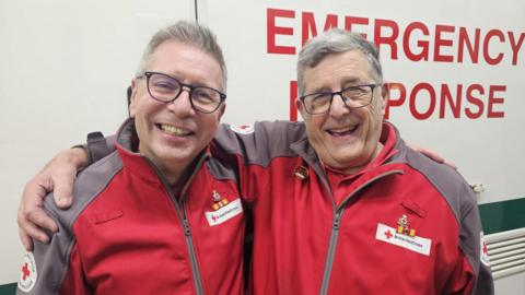 Andrew and Steve are smiling at the camera with their arms around one another. they both have short grey hear and are wearing black rimmed glasses. They are wearing matching red cross jackets. behind them is a vehicle with the words emergency response on.