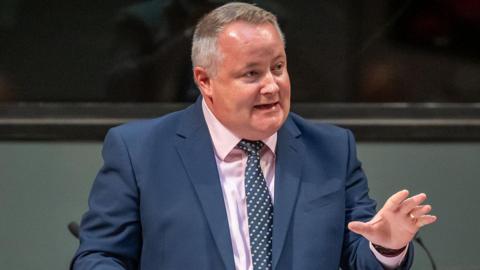Darren Millar gesturing with one hand, wearing a dark suit, pink shirt and a blue tie