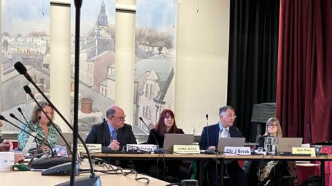 Councillors sit around a table in front of individual name cards. Some are speaking into table-top microphones.