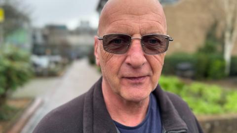 Dean Wedge wearing shaded glasses and a grey zipped fleece standing outside a hospital building