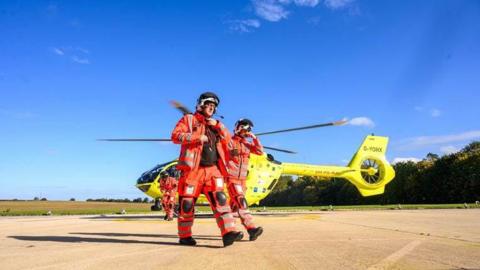 Two people wearing high‑visibility orange protective flight suits and helmets walking across a concrete landing area. Behind them is a yellow helicopter with its rotor blades visible and the registration marking “G‑YORK” clearly readable on the tail section. The setting appears to be an open airfield or helipad, with flat paved ground, grassy fields in the distance, and a clear blue sky overhead. 