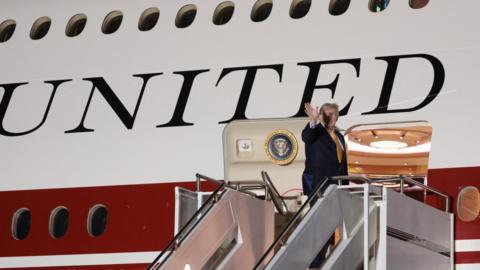 President Donald Trump is standing at the top of a set of steps leading to the door of a large plane. He is wearing a blue suit and is waving.
