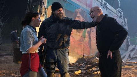 Millie Bobby Brown and David Harbour in character as Eleven and Jim Hopper. Eleven wears red shorts over grey leggings and a grey jumper. Jim Hopper wears a black leather jacket, a black beanie hat and dark trousers.They are speaking to director Frank Darabont on the set of Stranger Things. There is a vintage advertisement posted in the background with debrief on the ground. 