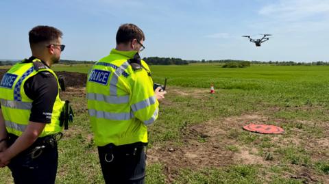 Two male police officers in hi-vis uniforms operate an aerial drone from a small landing pad in a field. It is a bright sunny day and one of the officers wears sunglasses.