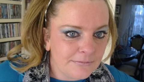 Jodey Whiting is looking towards the camera. She has blonde shoulder-length hair with a silver hairband and is wearing a a silver coloured scarf and a light blue jumper