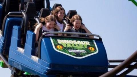 Three girls screaming in pleasure as they enjoy a roller-coaster ride. Their blue roller-coaster car is branded "Dinosaur Valley - Wicksteed Park"