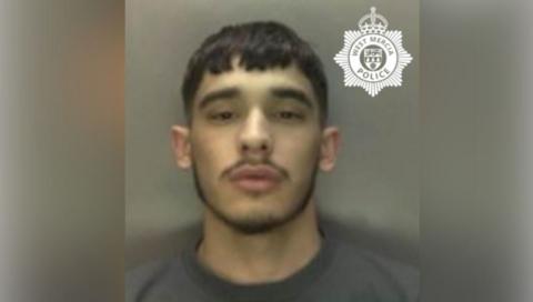 A man with shirt black hair with a black moustache is pictured in front of a neutral background.