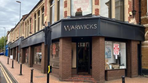 The outside of Warwicks now. It has closing down sales in the window, clothes in the main windows, and the front is made of brick, with a metal frontage for the sign in black. Bollards are outside the shop. There are steps up to the building. 