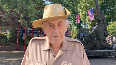 David Shopland looks at the camera with a serious expression. He wears a pale beige military shirt and a distinctive pale yellow hat with a wide rim and a military badge on the front. Behind him a camouflaged green military vehicle can be seen, as well as a Union Jack and the US flag hanging from a tree. 