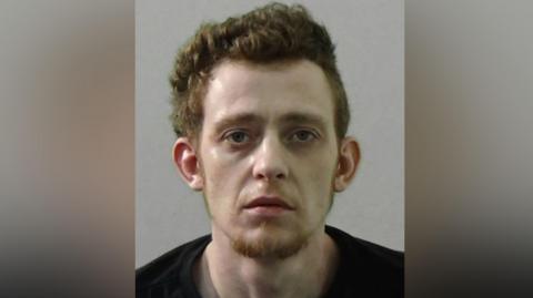 Mugshot of Reece Galbraith. He has thick ginger hair and a ginger goatee and a very pale face. He is wearing a black t shirt
