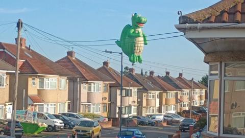 A large dragon-shaped hot air balloon is flying over residential streets with cars lined up.