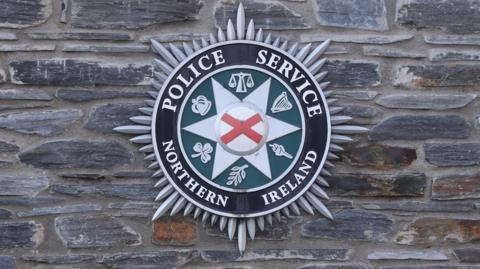 A PSNI badge is on a brick wall. The badge has the words 'Police Service of Northern Ireland' in white writing with a red cross in the middle and harps, scales, crowns and leaves on green background.