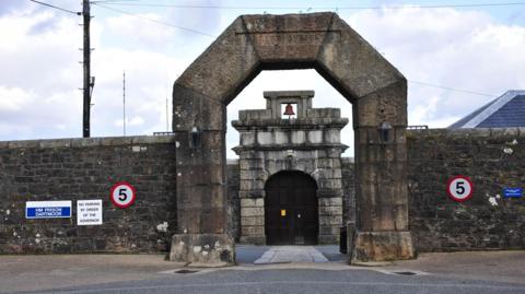 The entrance to HMP Dartmoor in Devon. It consists of a large stone arch and a large stone wall. Another archway with a door in it can be seen through the other arch.