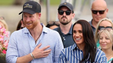 Harry and Meghan seen walking together in Sydney harbour in April. He is wearing a blue shirt and a cap, she is wearing a striped blue shirt.
