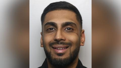 A police mugshot of Uzair Hamid. He has short dark hair and dark facial hair.