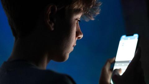 A teenage boy looks at an iPhone screen displaying various social media apps. It is dark in the room.