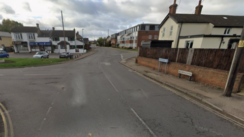 Greenfield Road leading into Flitwick, with houses and small shops on either side of the road and double yellow lines.