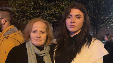 Two women standing next to her, one has shoulder length light brown hair and is wearing a black coat with a grey scarf. The woman on the right has long dark brown hair and is wearing a black top with a beige coat.