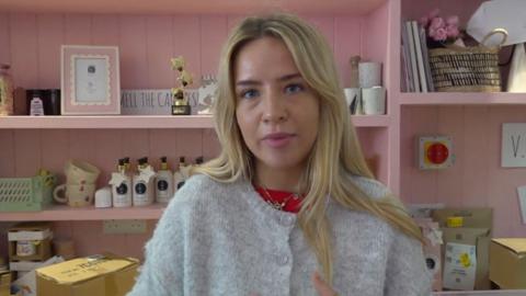 Promise Fulstow is a shop assistant at gift shop, U Melt Me, and is pictured behind the counter next to gifts and packaging and has blonde hair and wears a wool cardigan. 